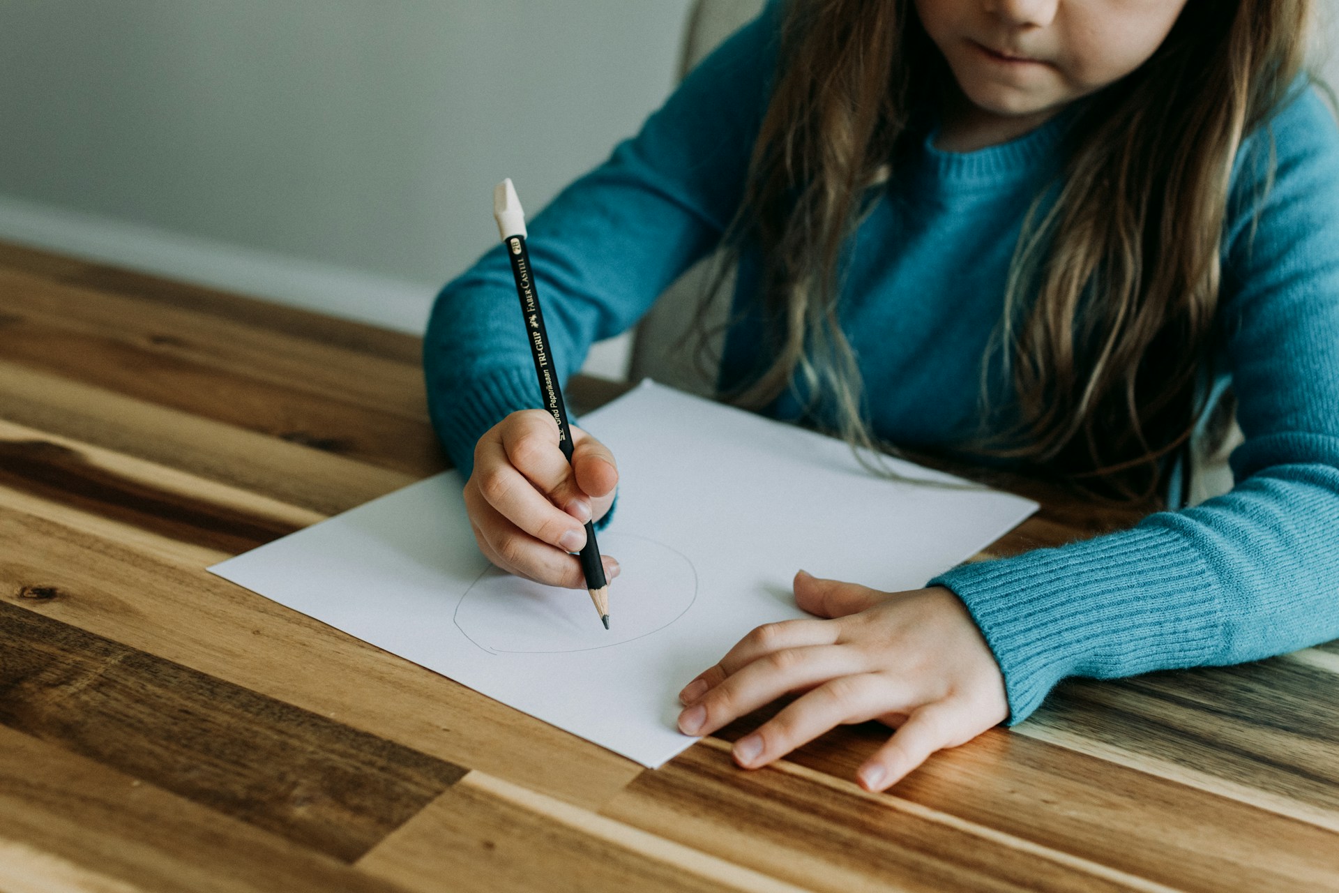 A young child in a blue sweater drawing a circle on a white sheet of paper on a wooden table