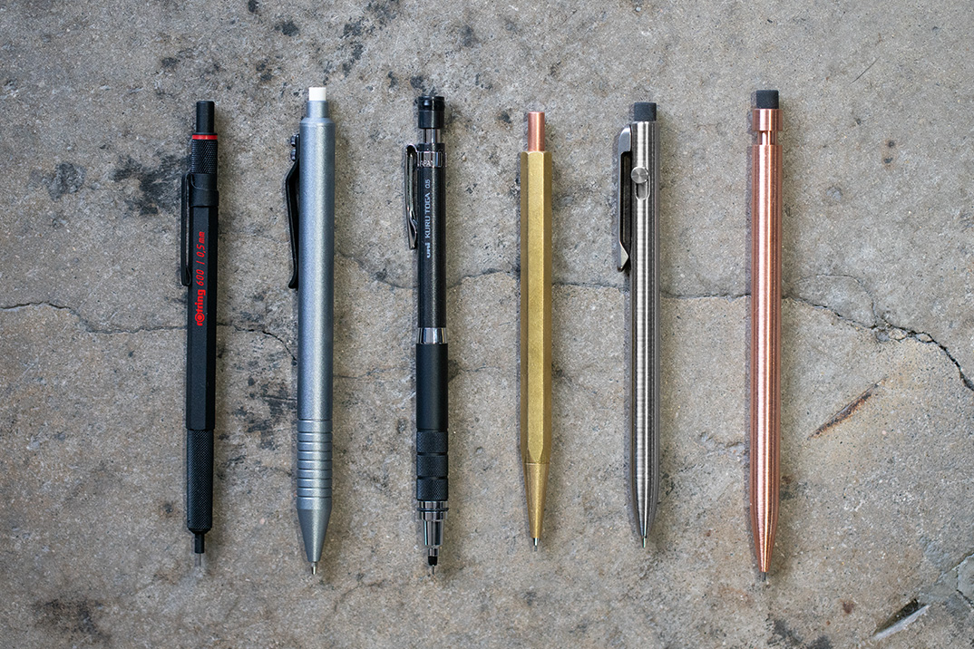 Top-down view of six professional mechanical and ballpoint pens in various metal finishes on a concrete surface