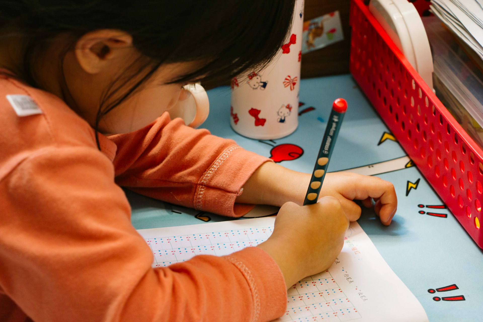 Close-up of a child in an orange shirt practicing writing on a worksheet with a dotted grid at a desk